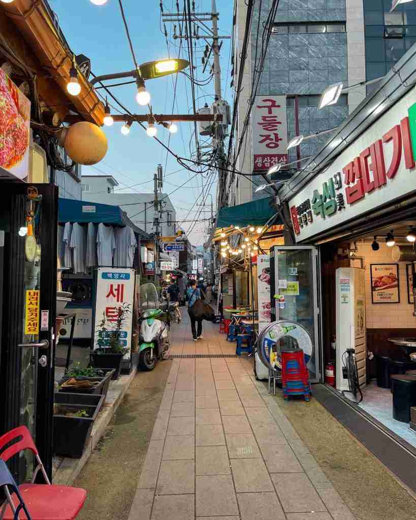 A bustling alleyway leading to Ikseondong Hanok Village, lined with small shops, restaurants, and glowing string lights. The hidden nature of the hanok village is hinted at by this narrow passage, offering a blend of modern storefronts and traditional charm.
