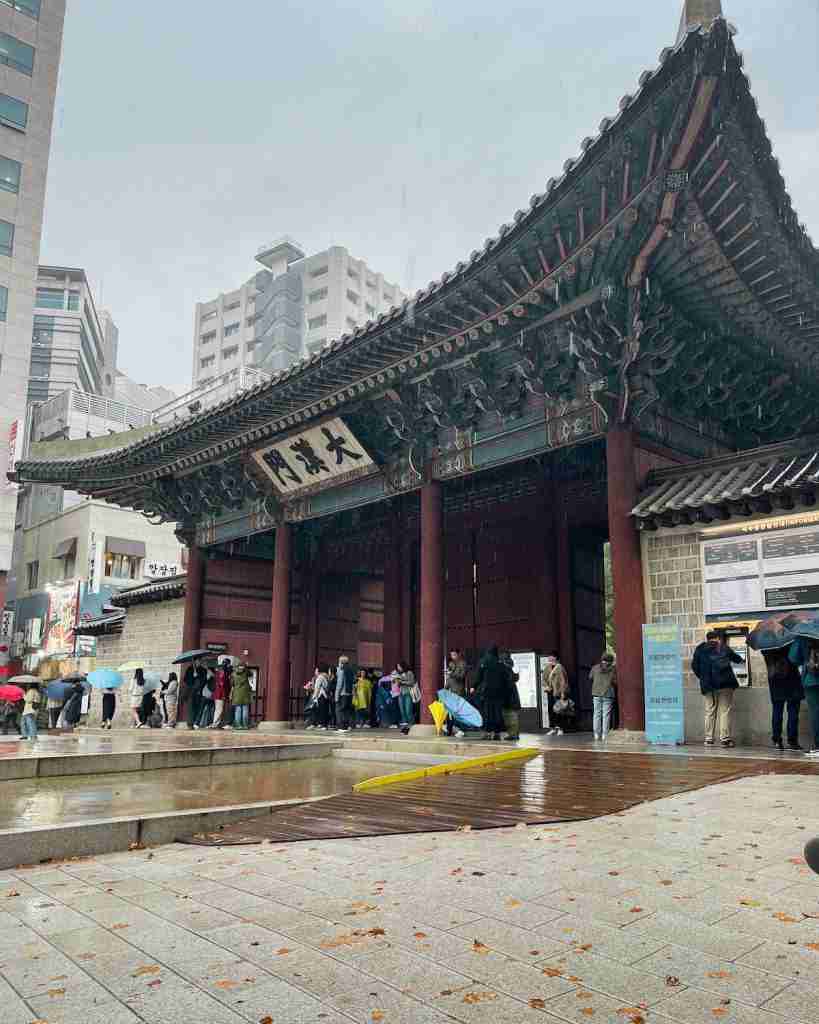 Daehanmun Gate of Deoksugung Palace in the rain, with brown autumn leaves fallen onto the ground in front. Tourists can be seen lining up outside the gate, holding umbrellas.