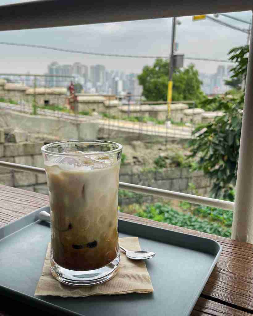 A refreshing iced coffee on a table, overlooking the Seoul City Wall and a distant view of the city skyline. A cozy moment captured in Ihwa Mural Village.