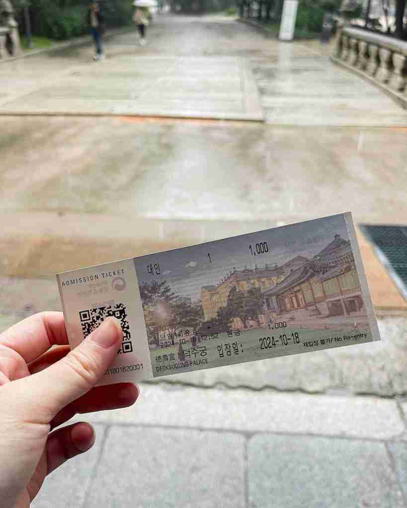 A hand holding a ticket to enter Deoksugung Palace. The tickets is a horizontal rectangle piece of paper with a QR code on the left side and a photo of a Western-style palace building on the right side.