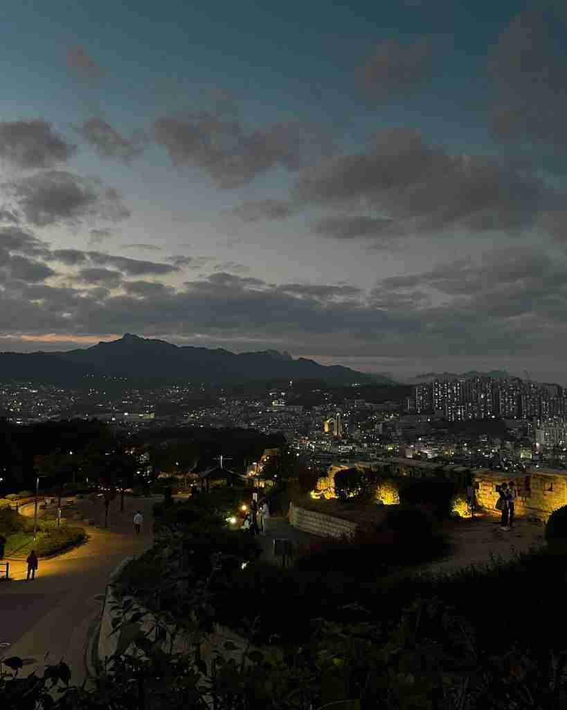 A serene evening view from Naksan Park, with softly lit pathways, stone walls, and benches overlooking the city of Seoul. The distant skyline and silhouetted mountains are set against a dramatic, cloudy twilight sky.