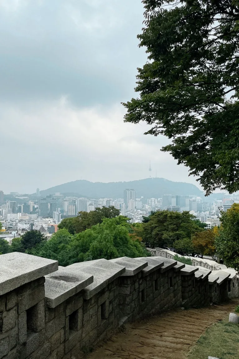A scenic view of Seoul, South Korea, from a historic stone wall pathway on a hillside. The city skyline, featuring modern high-rise buildings, stretches below with the iconic N Seoul Tower visible in the distance atop Namsan Mountain. Lush green trees frame the scene, creating a serene contrast between nature and urban life, perfect for exploring during a short 2-day visit to Seoul