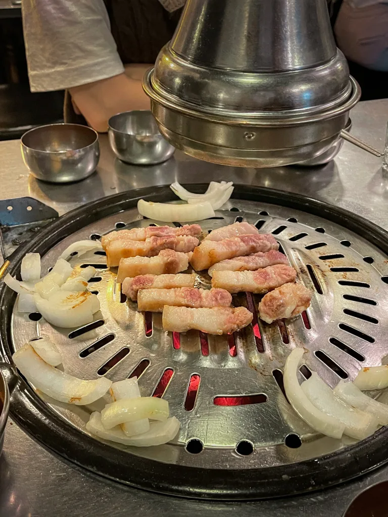 Thick slices of pork belly (samgyeopsal) being grilled alongside onions under a metal exhaust hood, a staple Korean dining setup.