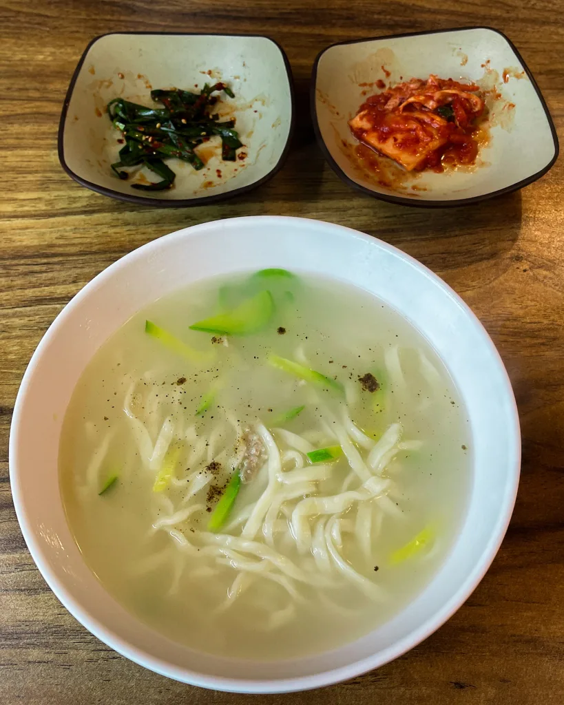 Steaming bowl of Kalguksu, Korean hand-cut noodles in a mild broth, garnished with minced meat and green onions, served with kimchi and chives.