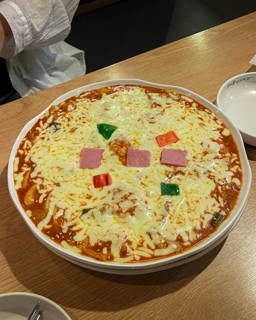 A large dish of ddeokbokki smothered in melted cheese with colorful toppings like ham cubes and bell peppers, a modern twist on a traditional snack usually shared with friends on a night out.