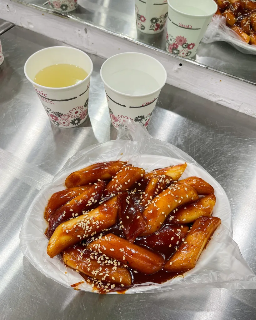 Spicy Korean rice cakes coated in gochujang sauce and sprinkled with sesame seeds, served in a plastic-lined plate with a cup of hot soup.