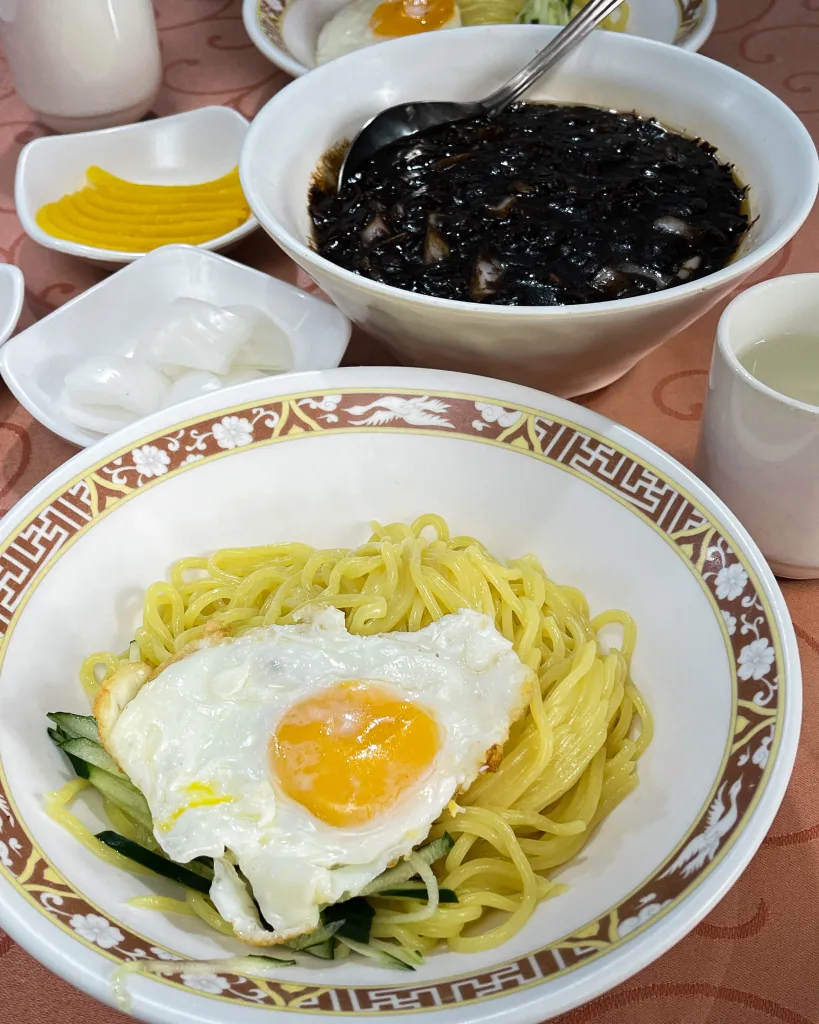 A bowl of rich jjajangmyeon with thick black bean sauce, accompanied by a plate of yellow pickled radish and cucumber noodles topped with a fried egg.