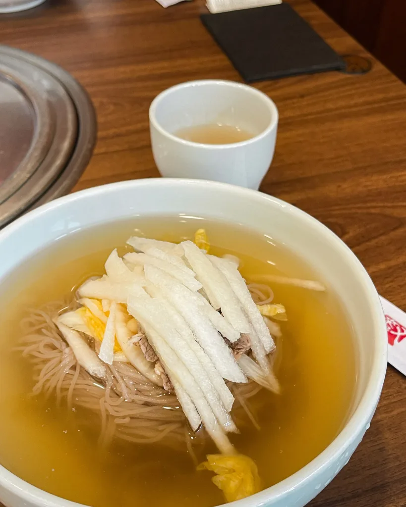 A bowl of mul naengmyeon (buckwheat noodles in icy cold broth) topped with pear slices and yellow pickled radish, refreshing and unique