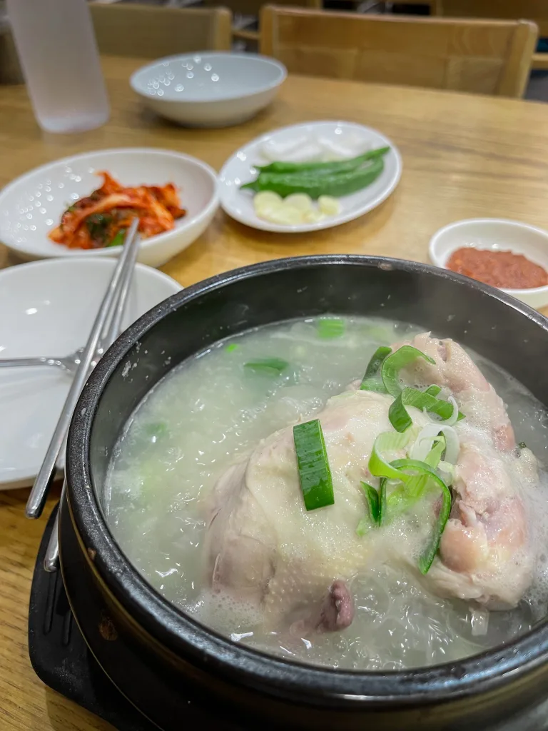 Boiling stone pot of Samgyetang, a whole stuffed chicken simmered in ginseng broth, served with side dishes like garlic, peppers, and kimchi. A traditional healing meal and cultural staple in what to eat in Seoul.