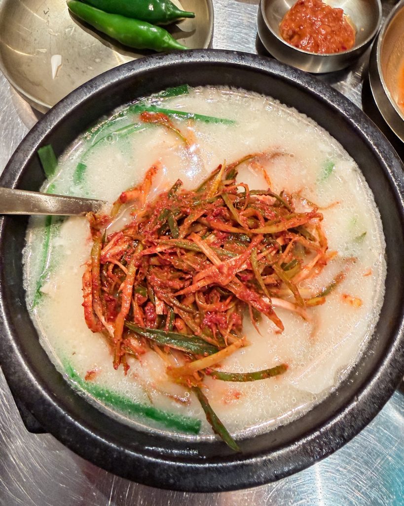 Steaming bowl of pork gukbap is topped with spicy seasoned spring onions and served with traditional Korean side dishes. This signature meal from Subyeon Chwego is a favorite place to try Busan-style pork soup.