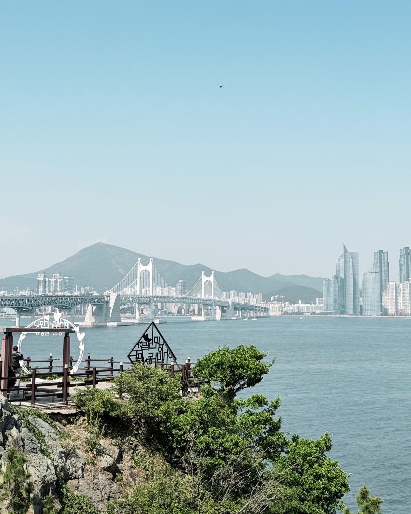 View from Igidae Coastal Walk overlooking a rocky shoreline and green trees beside the sea. The Gwangan Bridge stretches across the water toward Busan's skyline with mountains in the background under a clear blue sky.