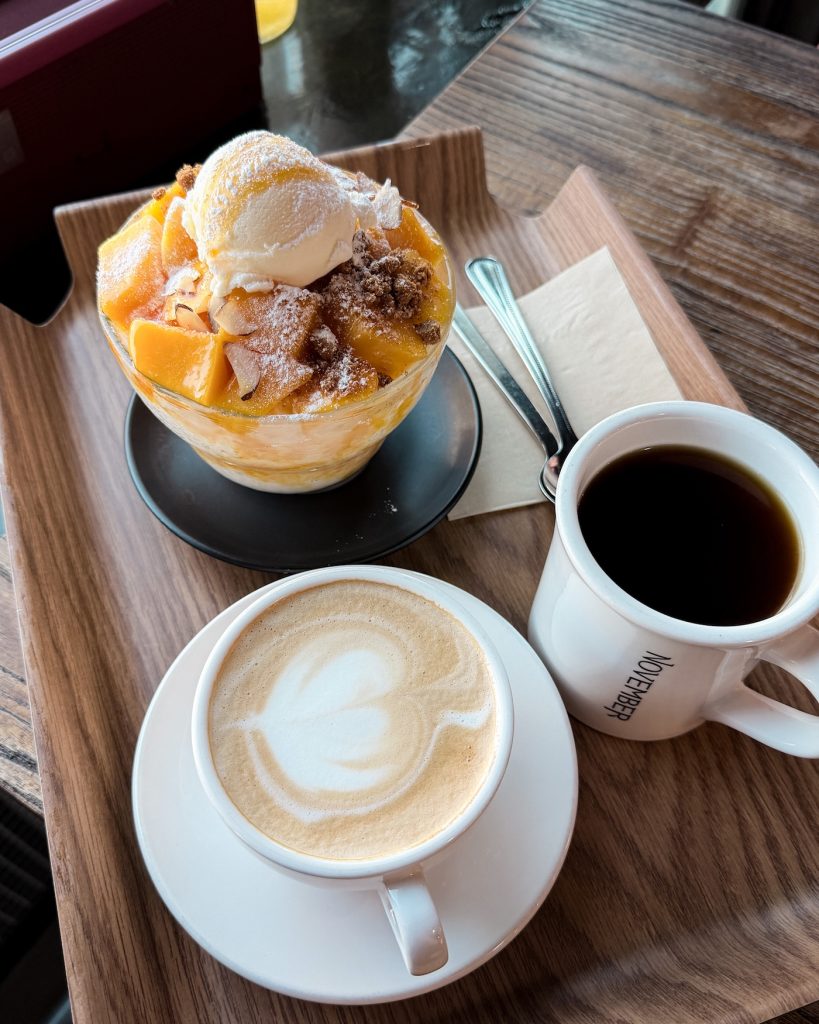 Mango bingsu topped with vanilla ice cream, fresh mango chunks, and almond slices served with a latte and black coffee on a wooden tray at November cafรฉ. This refreshing dessert and smooth coffee are recommended specialties at one of Gyeongju's best local cafรฉs.