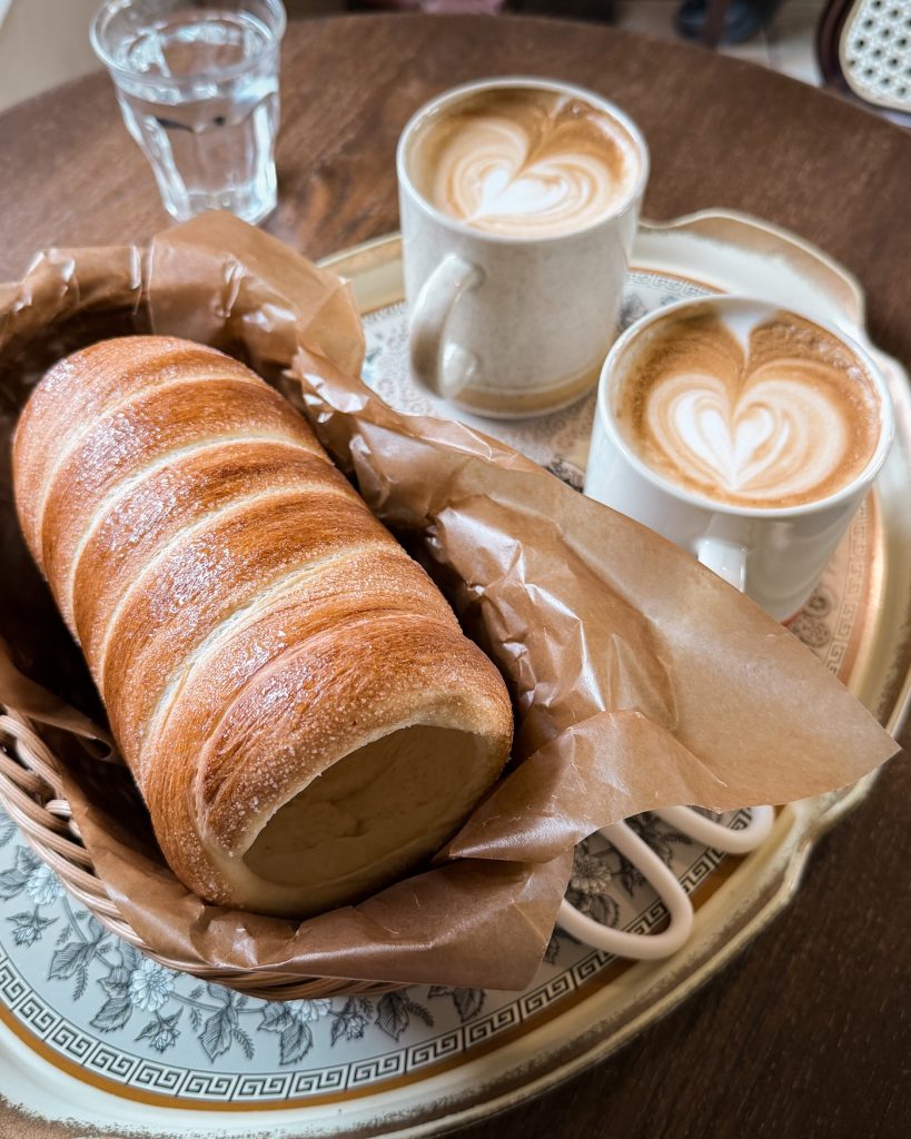 Freshly baked trdlo pastry rests on a wooden serving board beside two latte drinks at Cafรฉ Bohemia. The warm chimney cake makes a satisfying dessert while cafรฉ hopping around Oncheon-cheon.