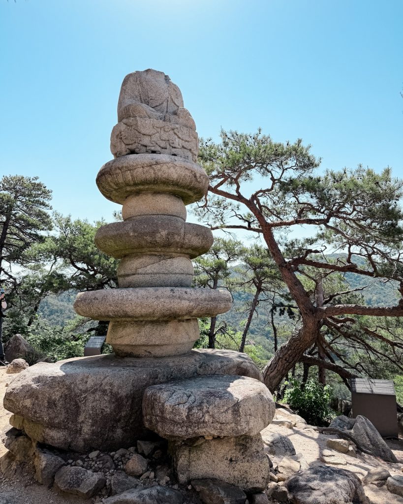 Weathered stone Buddha statue, with its head cut off, on Mount Namsan in Gyeongju overlooking forested hills beneath a clear blue sky.