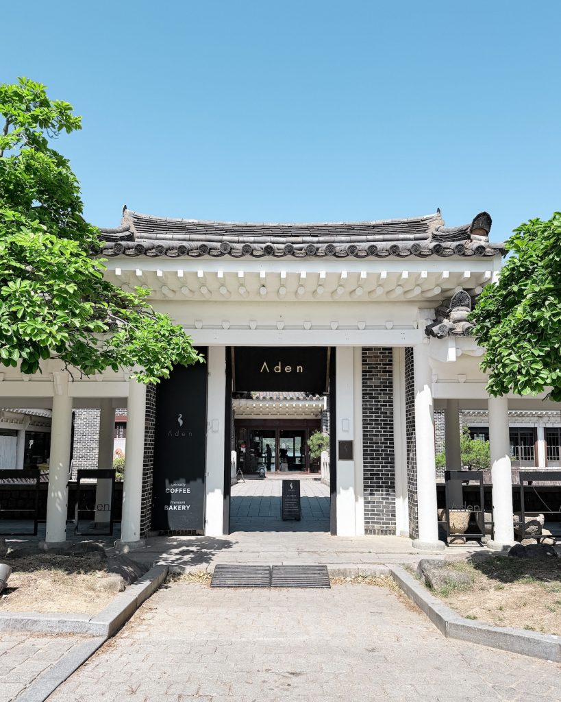 The hanok-style entrance to Cafe Aden at Bomun Lake, framed by leafy trees beneath a clear blue sky. Its traditional architecture makes it a popular stop before or after a walk around the lakeside.