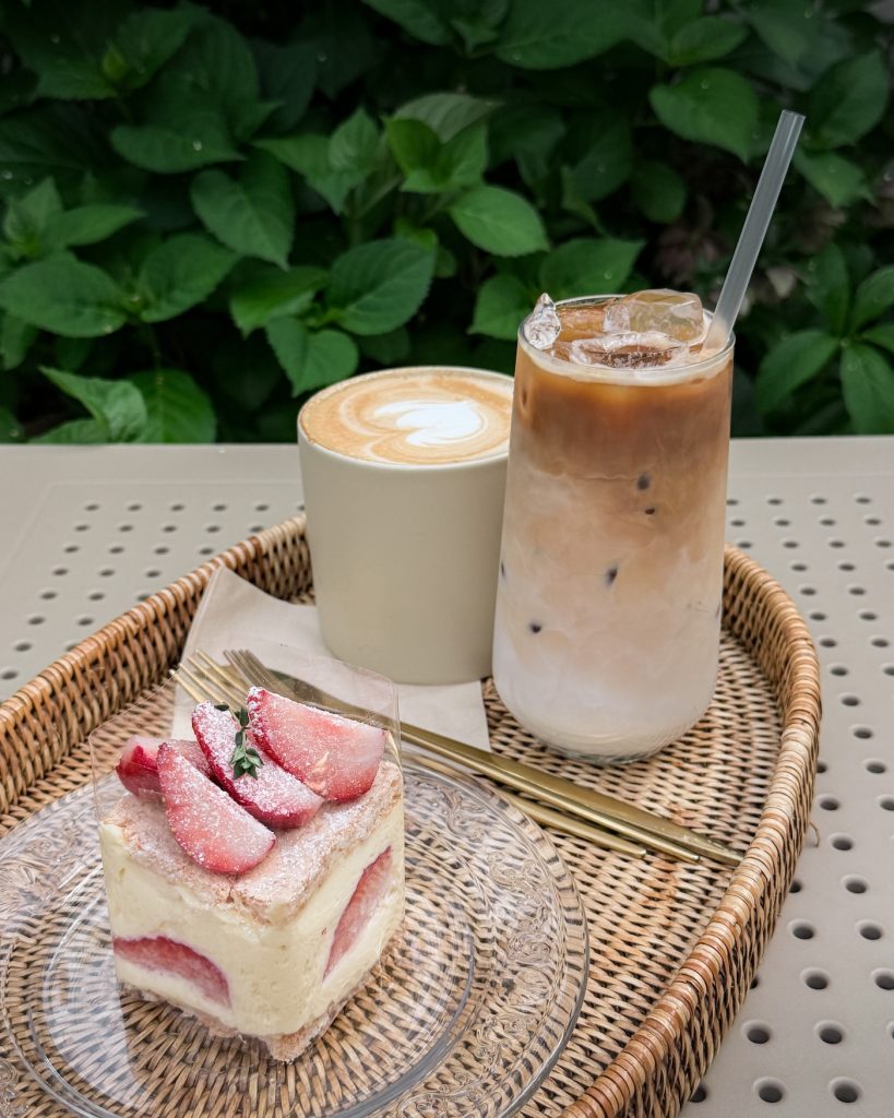 Slice of strawberry mascarpone cake and two iced lattes sit on a woven tray at Cheek to Cheek cafรฉ surrounded by greenery. This cozy neighborhood cafรฉ is a relaxing stop after exploring Oncheon-cheon.