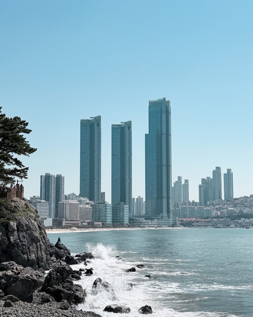Waves crash against rocky shoreline below the coastal cliffs at Dongbaekseom with Haeundae's modern skyscrapers rising across the bay. The trail is a nice alternative to Igidae for visitors with less time in the city.