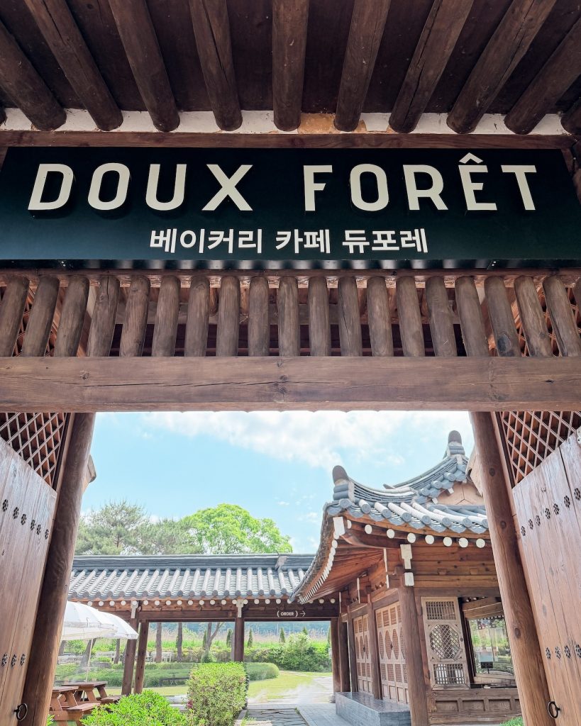 Wooden entrance gate with the sign "DOUX FORรT" and traditional hanok buildings visible beyond the courtyard. This bakery cafรฉ combines classic Korean architecture with spacious outdoor seating.