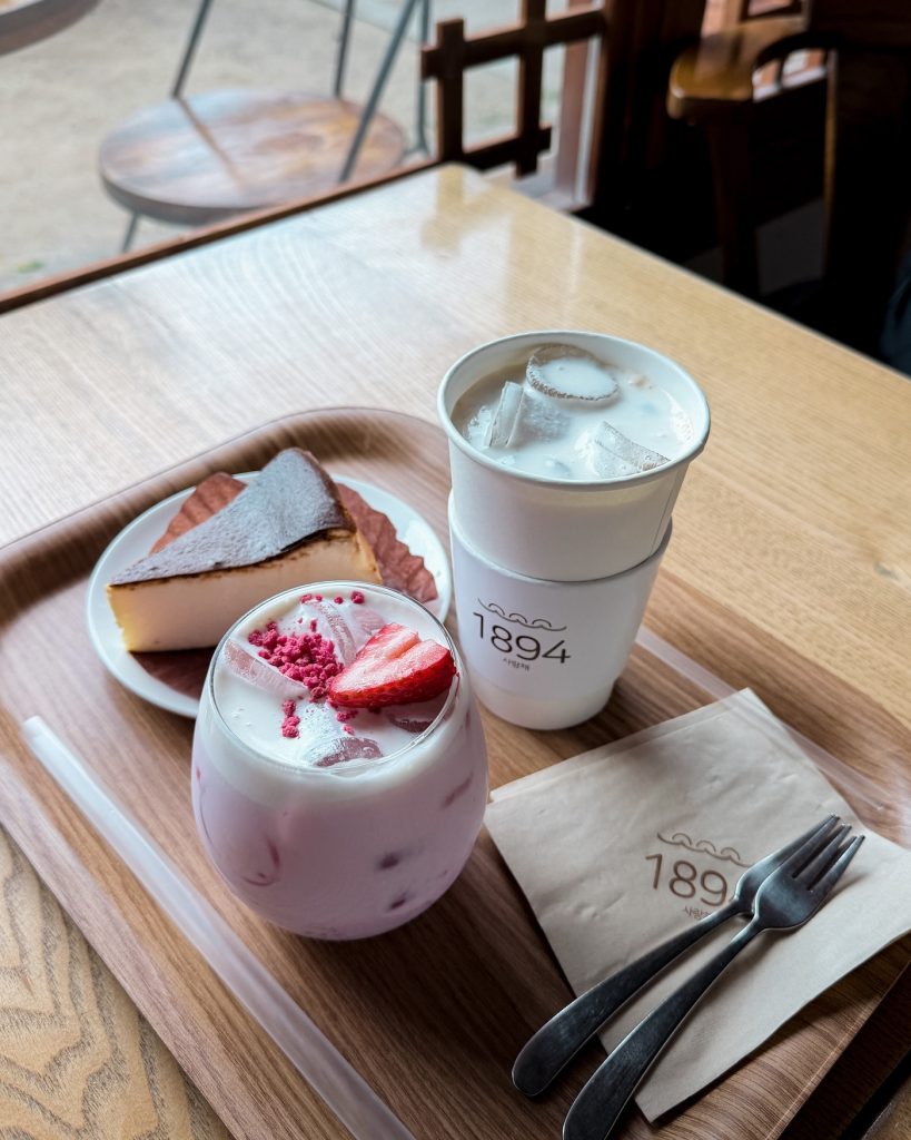 Slice of plain cheesecake, strawberry milk, and an iced vanilla latte served together on a wooden tray at Cafe 1894. These house recommendations make an easy afternoon stop after exploring central Gyeongju.