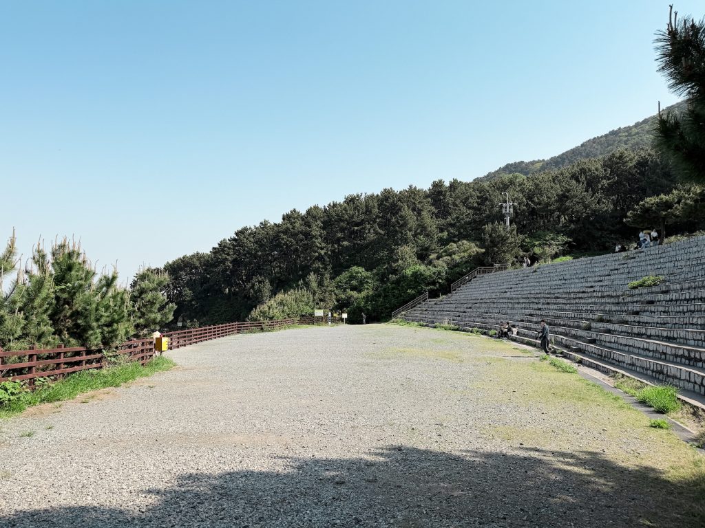 Stone seating terraces at Eoulmadang Amphitheater surrounded by pine forest inside Igidae Coastal Park. The open gravel performance area sits beside a wooden fence overlooking the coast.