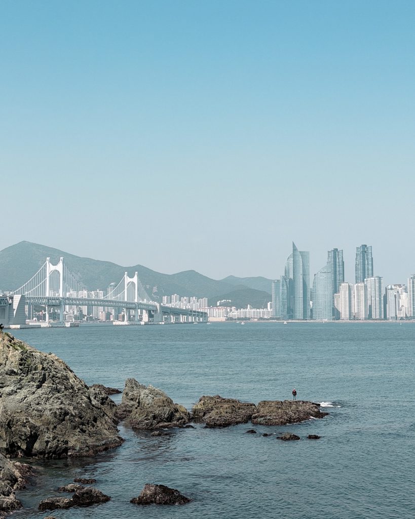 Lone fisherman standing on rocky outcrops in the sea below Gwangan Bridge and Busan's waterfront skyline. Calm blue water surrounds the rocks with mountains visible behind the city.