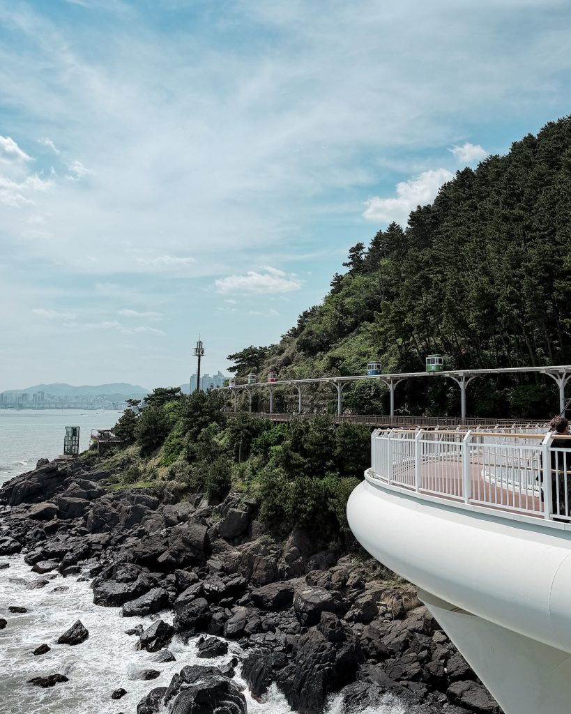 Haeundae Blueline Park Sky Capsules travel along an elevated coastal track above rocky cliffs and the sea. A white observation platform extends over the shoreline in the foreground.