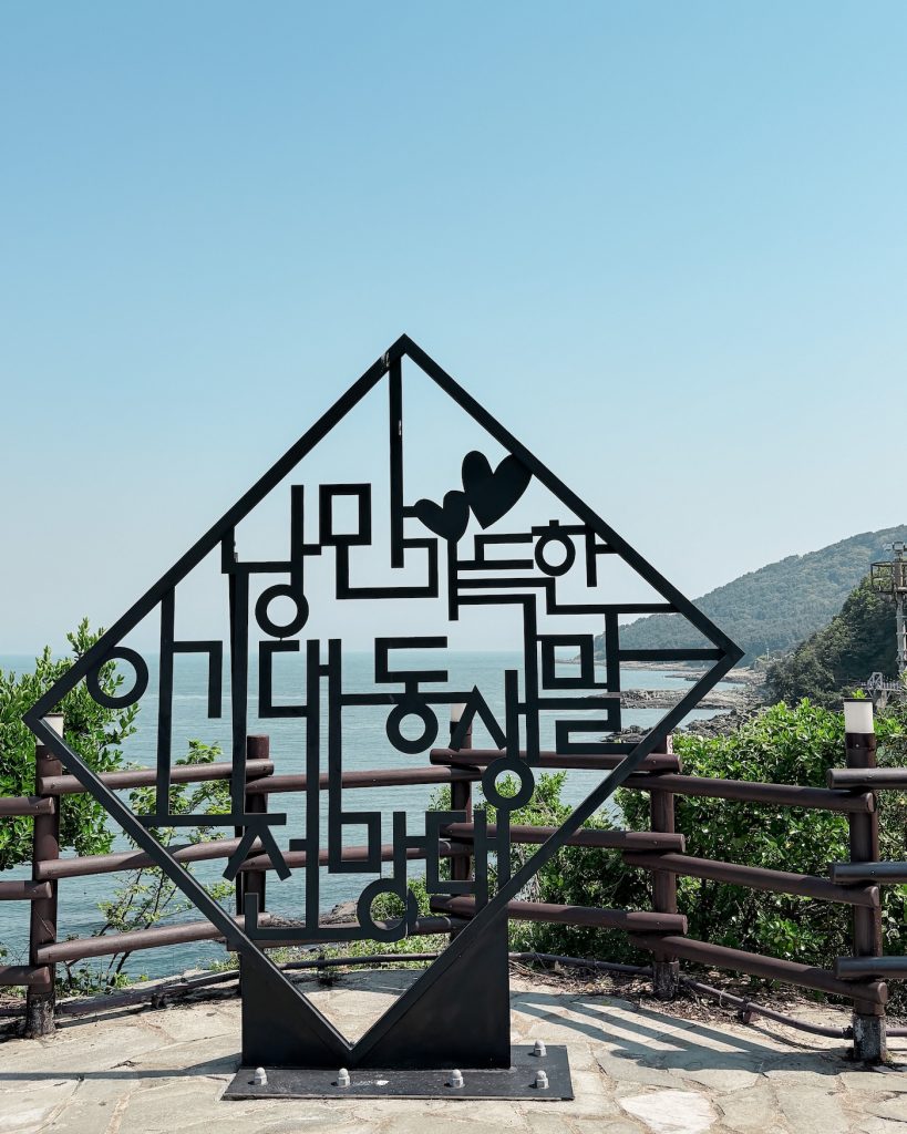 Black metal sculpture displaying a Korean phrase with heart shapes incorporated into the design. The sign stands at a coastal viewpoint on the Igidae Coastal Trail.