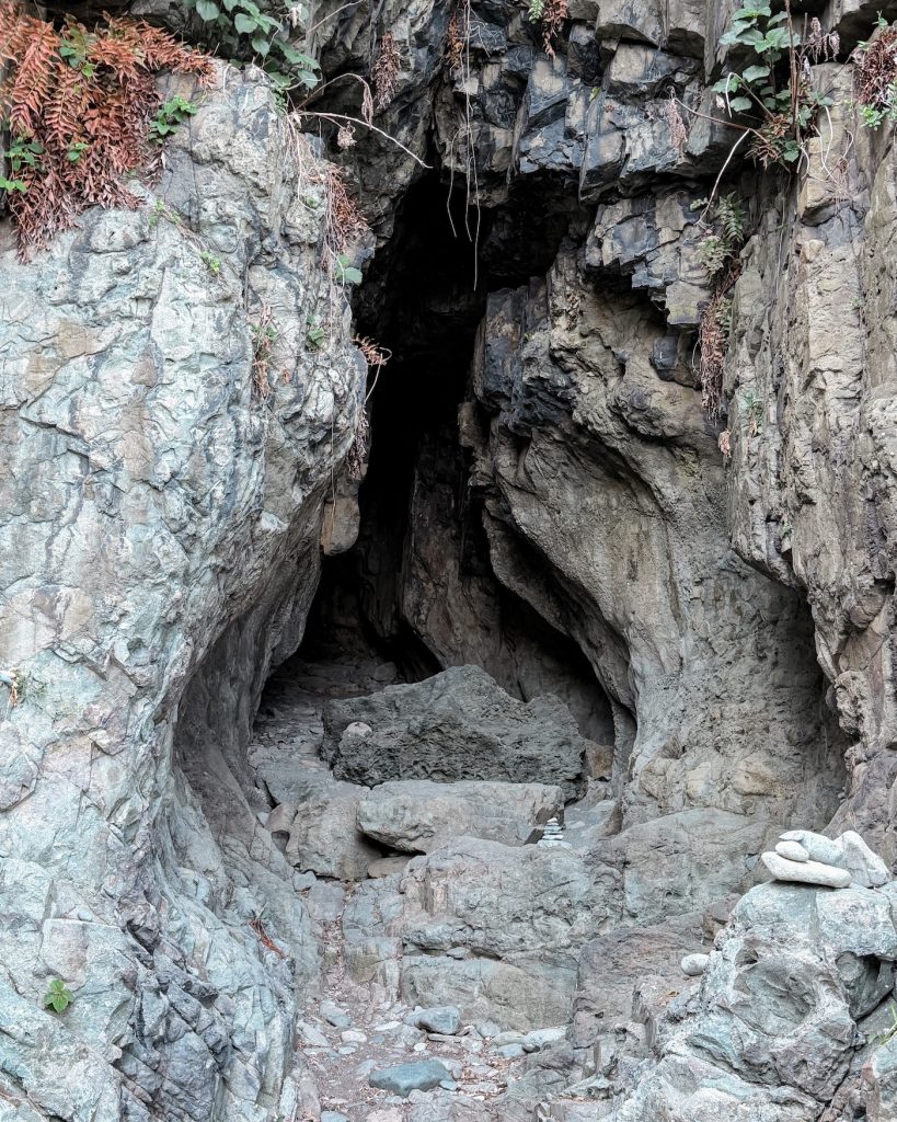 Narrow sea cave opening carved into a rocky cliff face along the Igidae coast. Large stone formations and weathered rock textures surround the dark entrance.