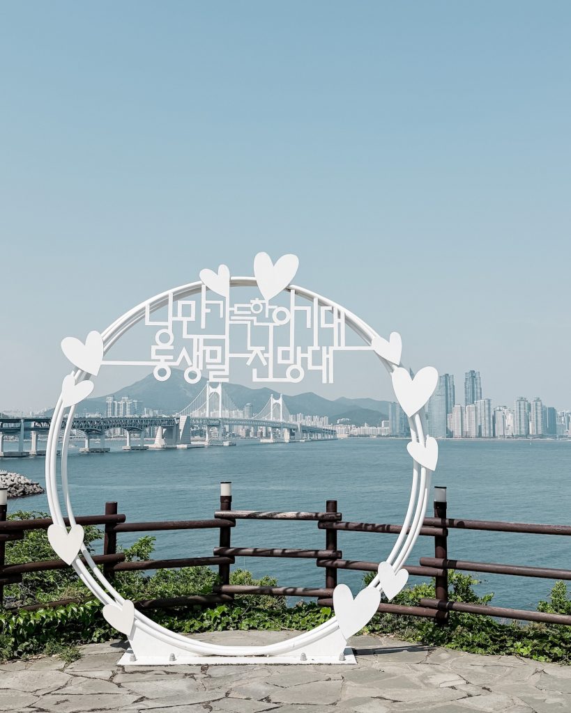 White circular photo frame decorated with heart shapes overlooking Gwangan Bridge and Busan's skyline across the bay. Korean text is integrated into the structure at the center of the viewing platform.