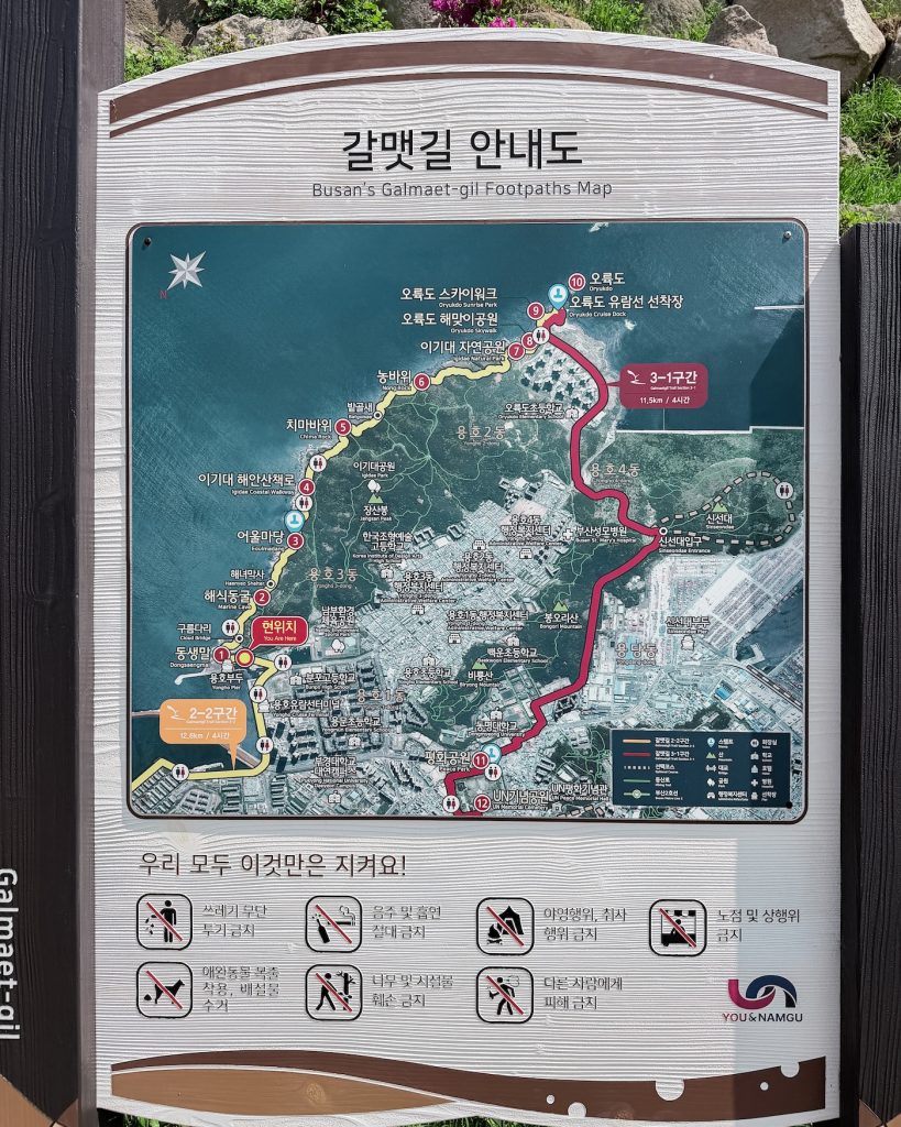 Trail information board titled "Busan's Galmaet-gil Footpaths Map" showing the coastal walking route through Igidae and Oryukdo. The sign includes a detailed map, trail sections, landmarks, and visitor guidelines.