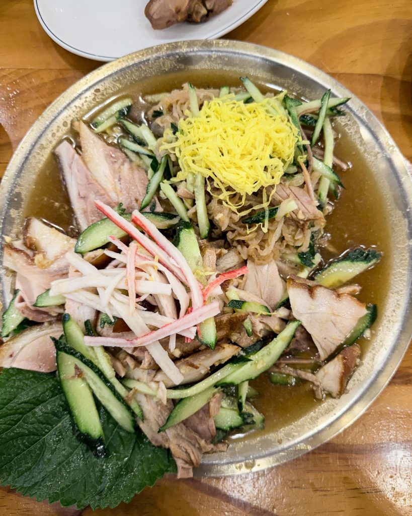 Chilled naengchae jokbal is served with sliced pork trotters, fresh cucumber, mustard dressing, shredded egg, and vegetables in a large metal bowl. This Busan specialty offers a refreshing twist on traditional jokbal.