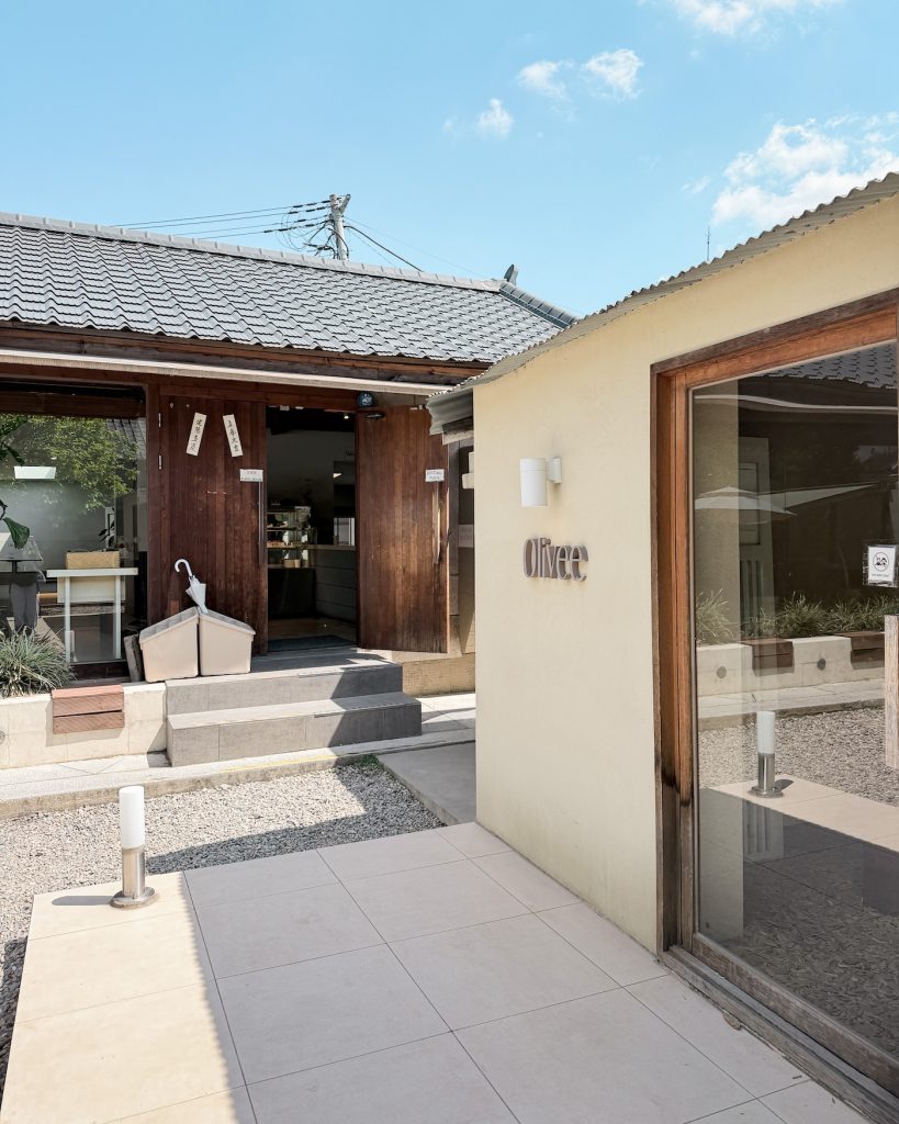 The entrance to Olivee cafรฉ features a minimalist cream colored exterior beside a restored hanok building with a small courtyard. The blend of modern and traditional architecture creates a peaceful atmosphere.