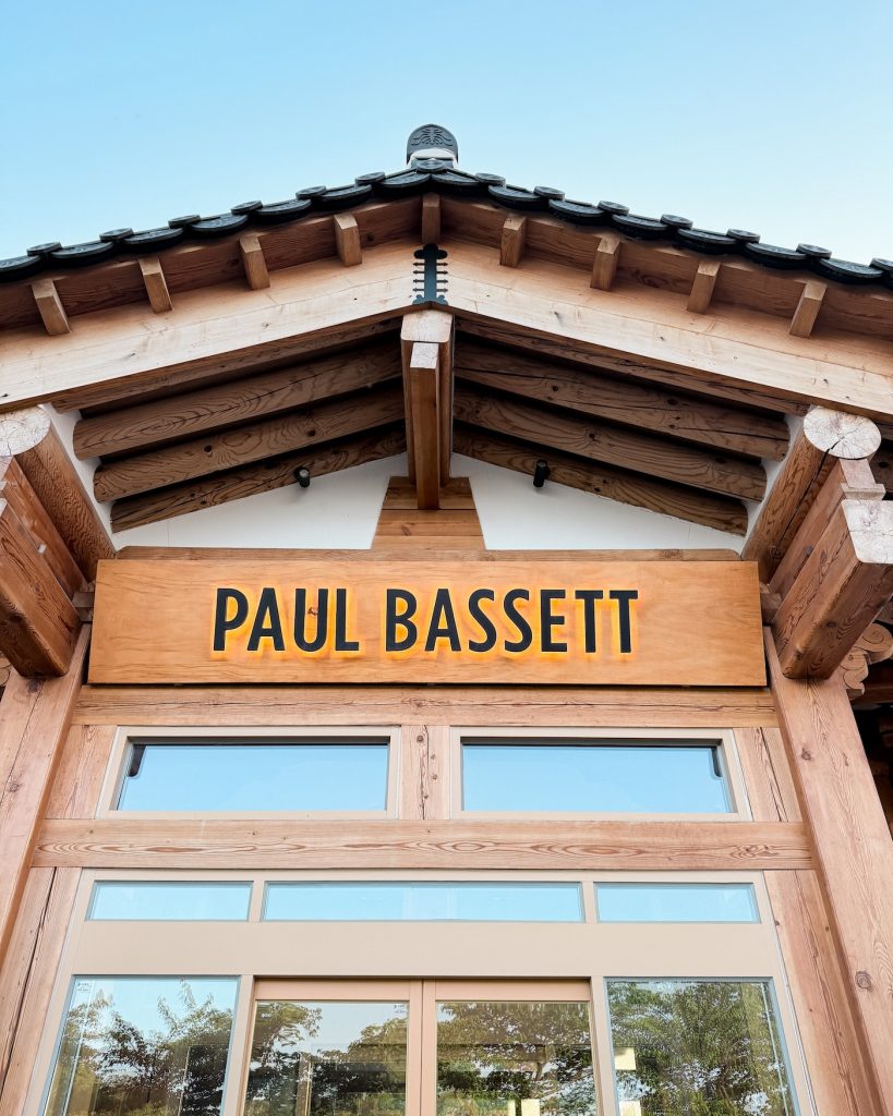 The wooden entrance to Paul Bassett features a large illuminated "PAUL BASSETT" sign beneath a traditional hanok style roof. The architecture combines Korean design with the cafรฉ's modern branding.
