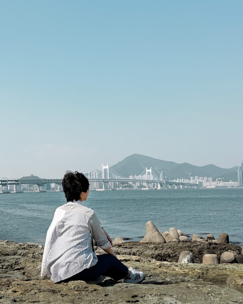 Petra, the author, seated on rocky shoreline at Igidae, looking across the water toward Gwangan Bridge with mountains and Busan's skyline in the background. The coastal viewpoint offers an expansive view of the city and sea.