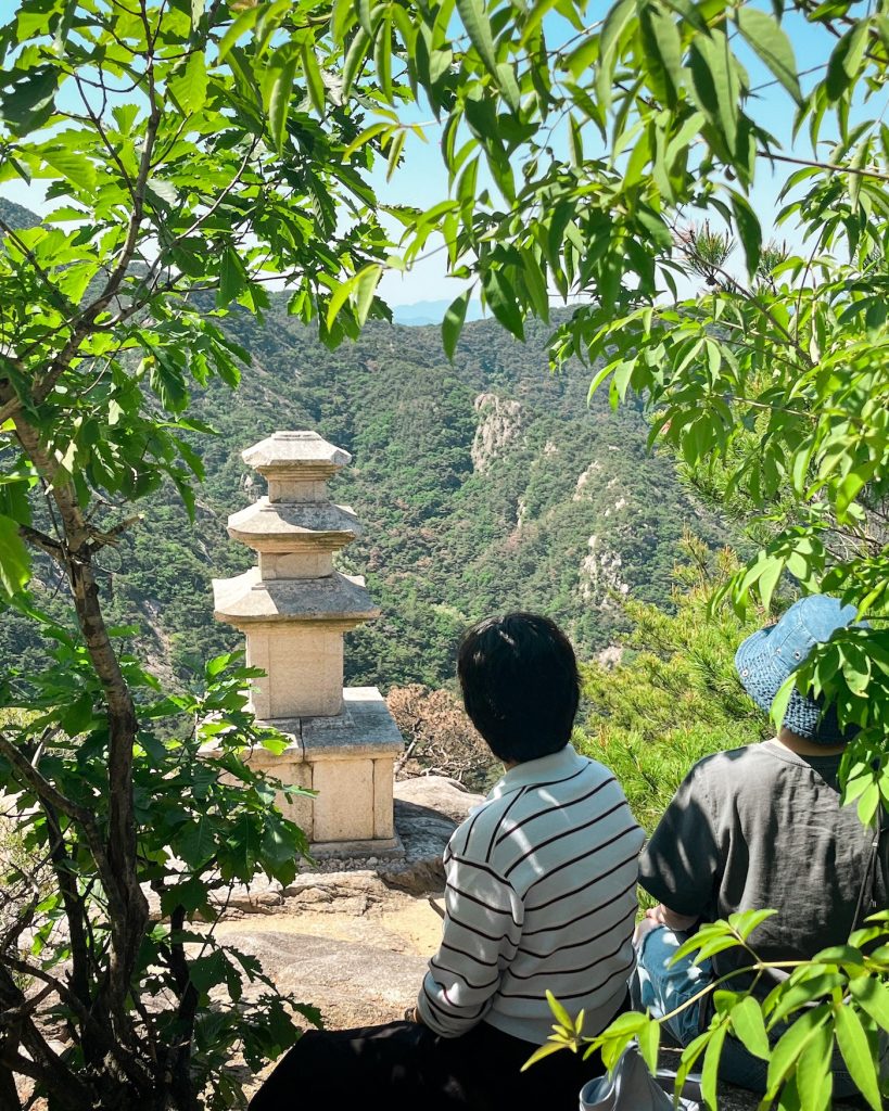 Two hikers sit beside the three tier stone pagoda at the Yongjangsa Temple Site on Mount Namsan in Gyeongju while looking out over the surrounding mountains.