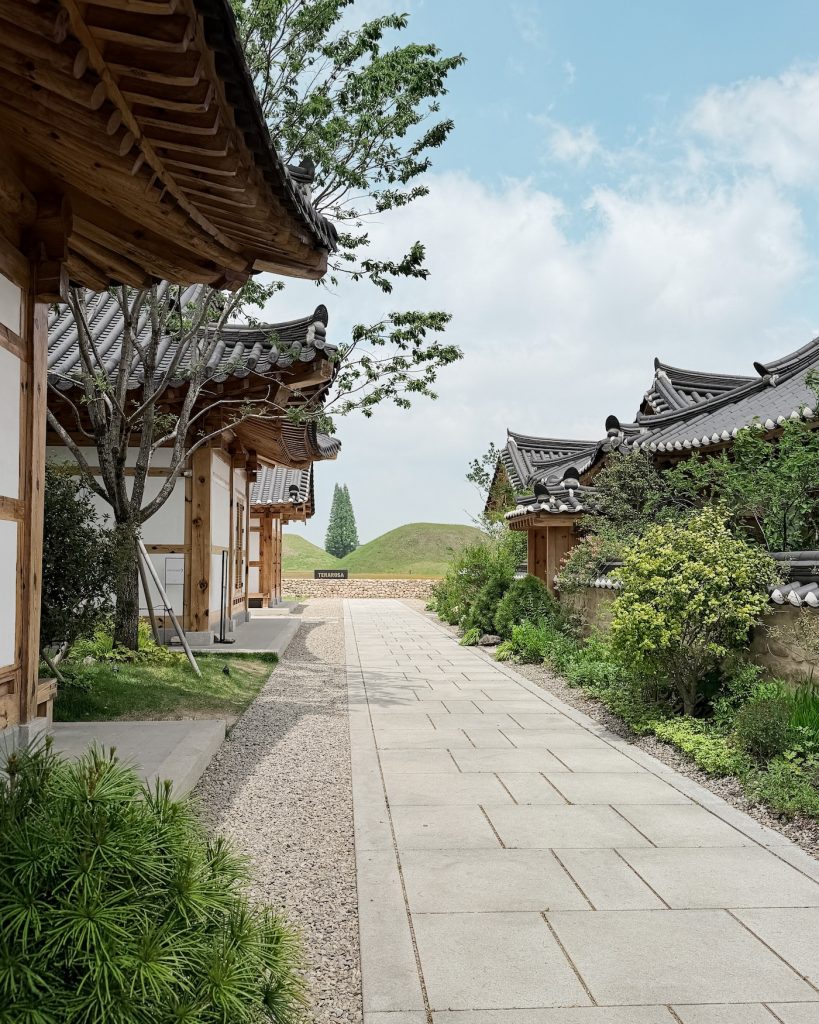 A stone pathway lined with traditional hanok buildings leads toward the TERAROSA grounds and grassy burial mounds in the distance. The peaceful setting blends Korean architecture with views of Gyeongju's historic landscape.