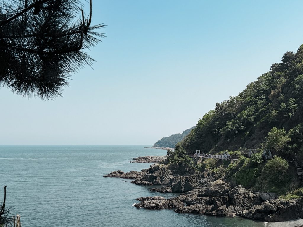 Rocky coastline and forested hills along the first part of Igidae Coastal Trail, stretched along the sea with pedestrian bridges built into the cliffs. Calm blue water borders the rugged shoreline under a clear sky.