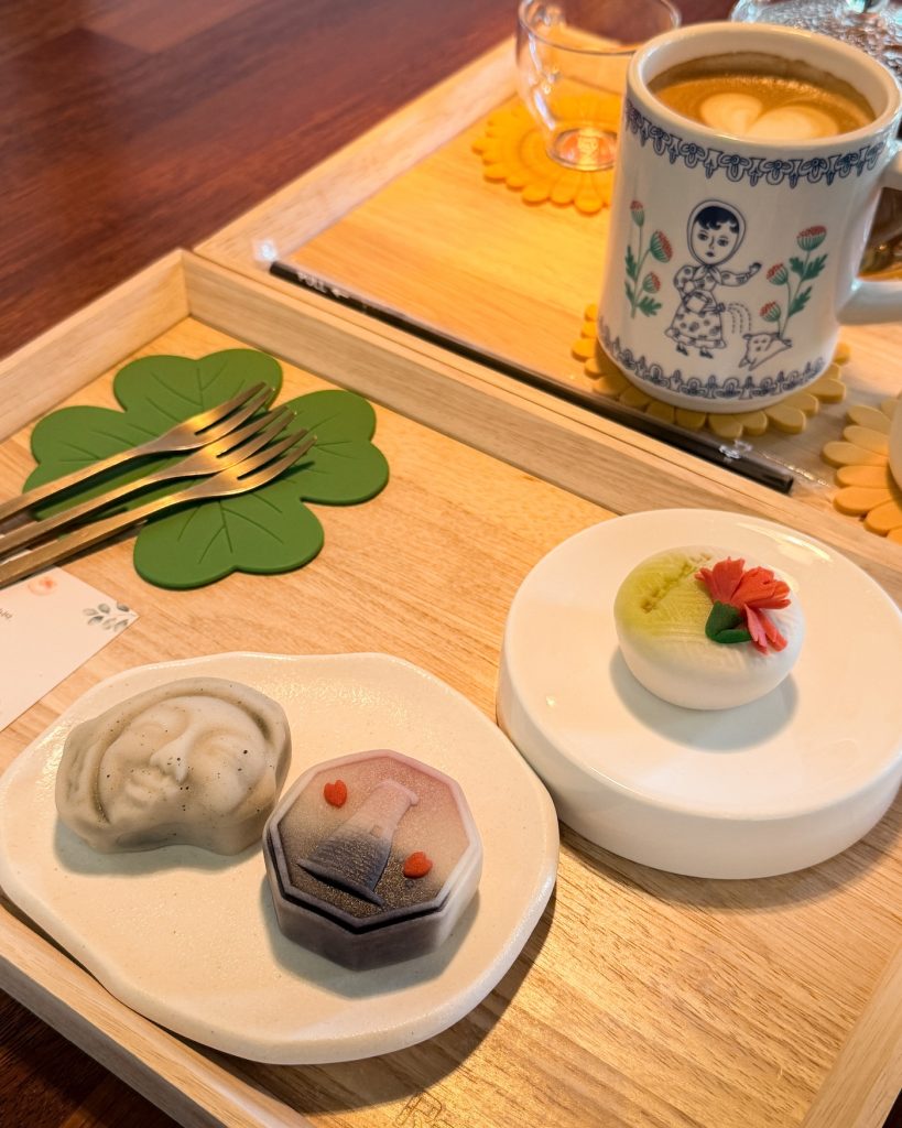 Three handcrafted wagashi sweets with delicate floral and artistic designs are served on small plates beside a latte in a decorative mug at Yunigarden Cafรฉ. The traditional Japanese style confections add a colorful and elegant touch to the cafรฉ experience.