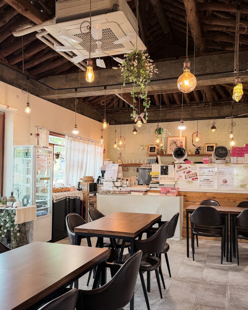 The cozy interior of Yunigarden Cafรฉ features exposed wooden ceiling beams, warm pendant lighting, and comfortable seating surrounding the service counter. The welcoming space blends rustic architecture with charming vintage inspired dรฉcor.