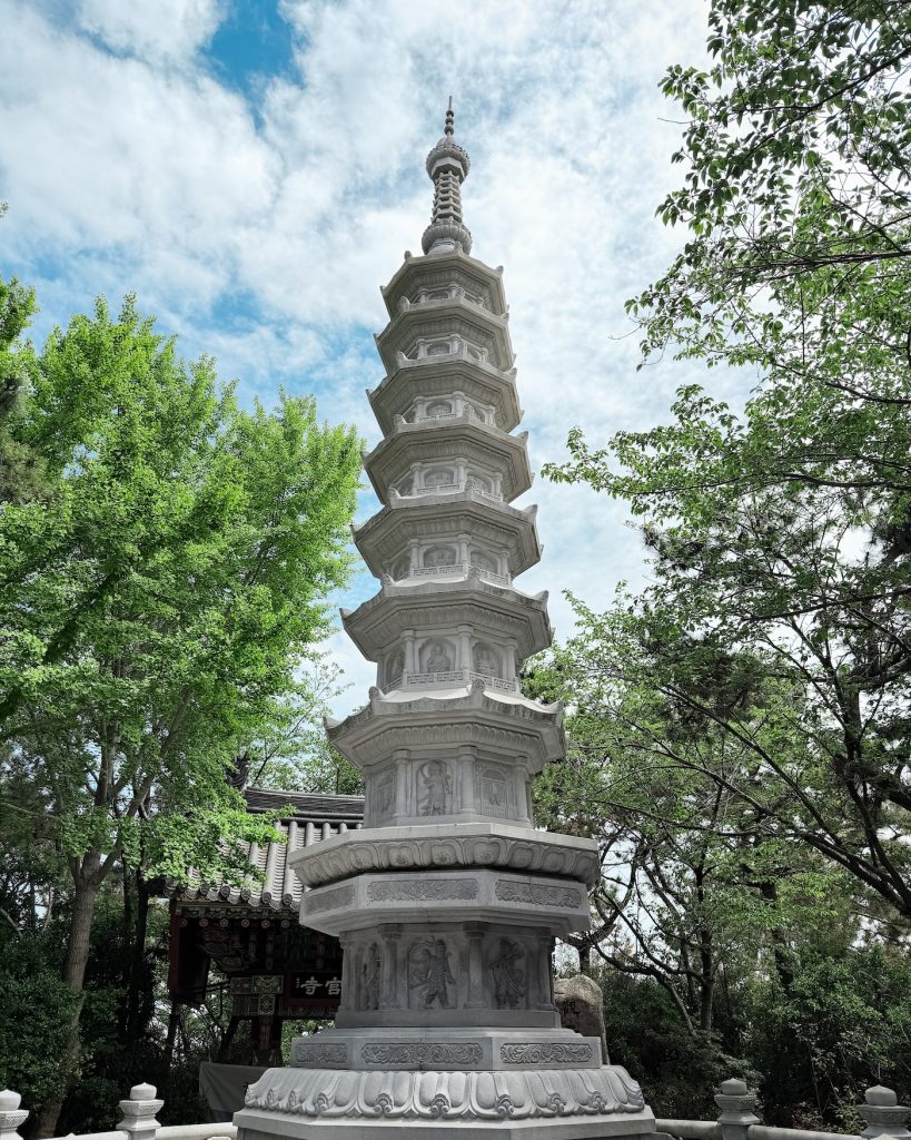 White stone pagoda rises above the grounds of Haedong Yonggungsa Temple, surrounded by lush green trees beneath a partly cloudy sky. The pagoda is one of the first landmarks visitors encounter while exploring Busan's famous seaside temple.