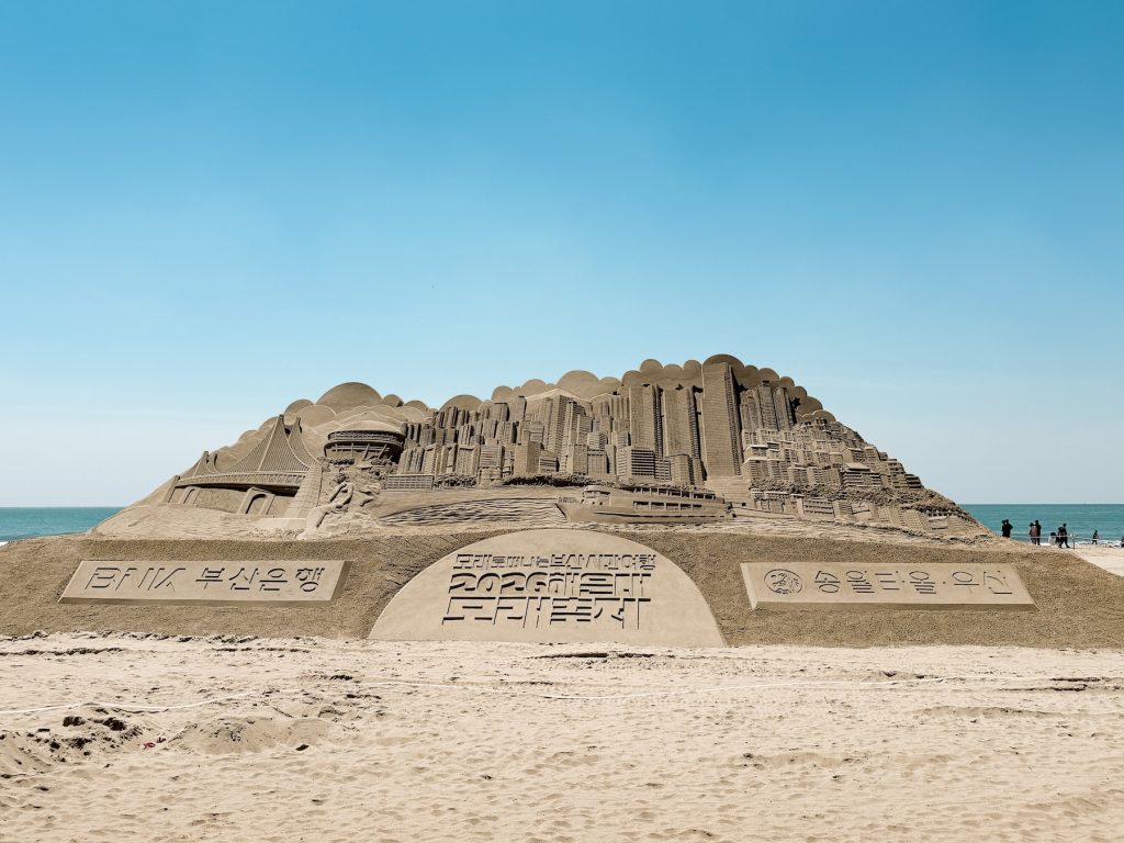 Massive sand sculpture depicting Busan landmarks stands on Haeundae Beach during the annual Sand Festival. The detailed carving showcases the festival's elaborate artwork along Korea's most famous urban beach.