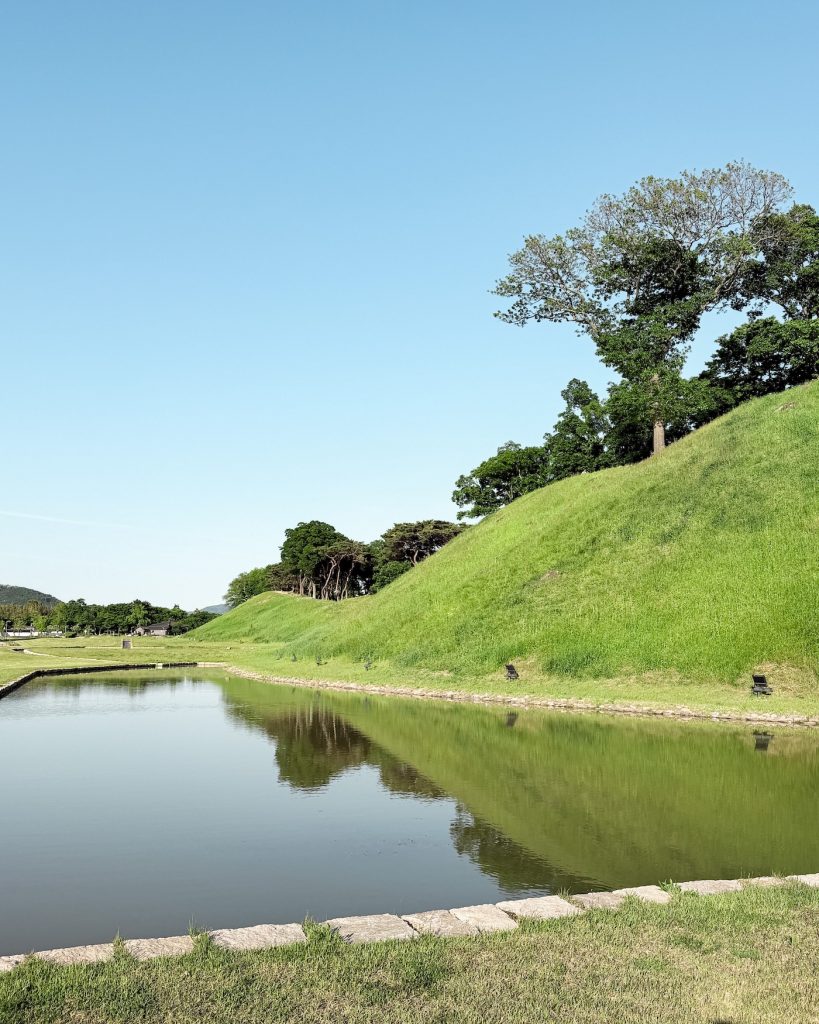 Grassy embankments of Wolseong Palace Grounds rise beside a calm moat on a sunny afternoon. The former Silla royal palace is now a peaceful park where visitors can stroll among ongoing archaeological excavations.