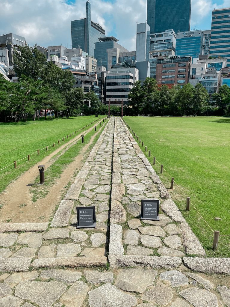 Stone spirit path crossing the lawn at Seolleung and Jeongneung with modern Gangnam buildings rising behind it. The path is traditionally reserved for spirits rather than visitors to the royal tombs.